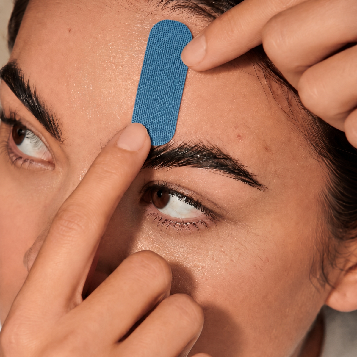 A close-up of a person applying a textured blue adhesive face tape strip to their forehead from HOLLI Face Tape infused with Hyaluronic Acid.